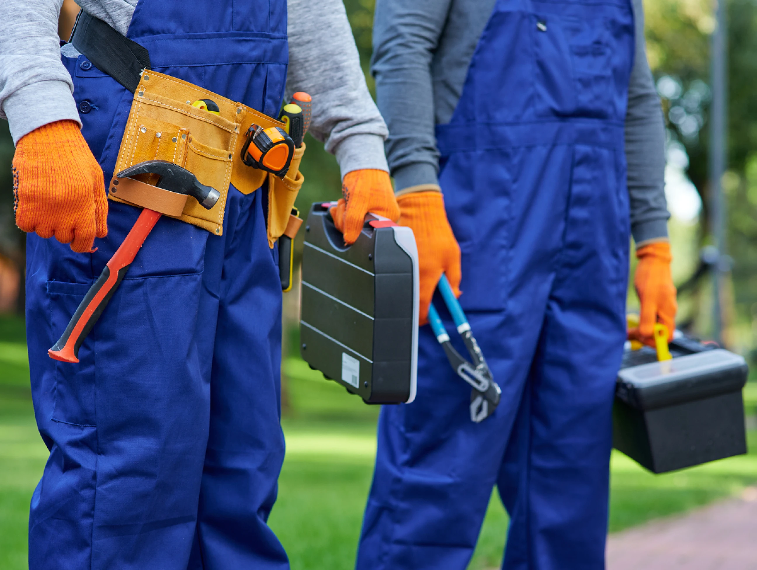 Two field service technicians in blue coveralls carrying tools and equipment outdoors.